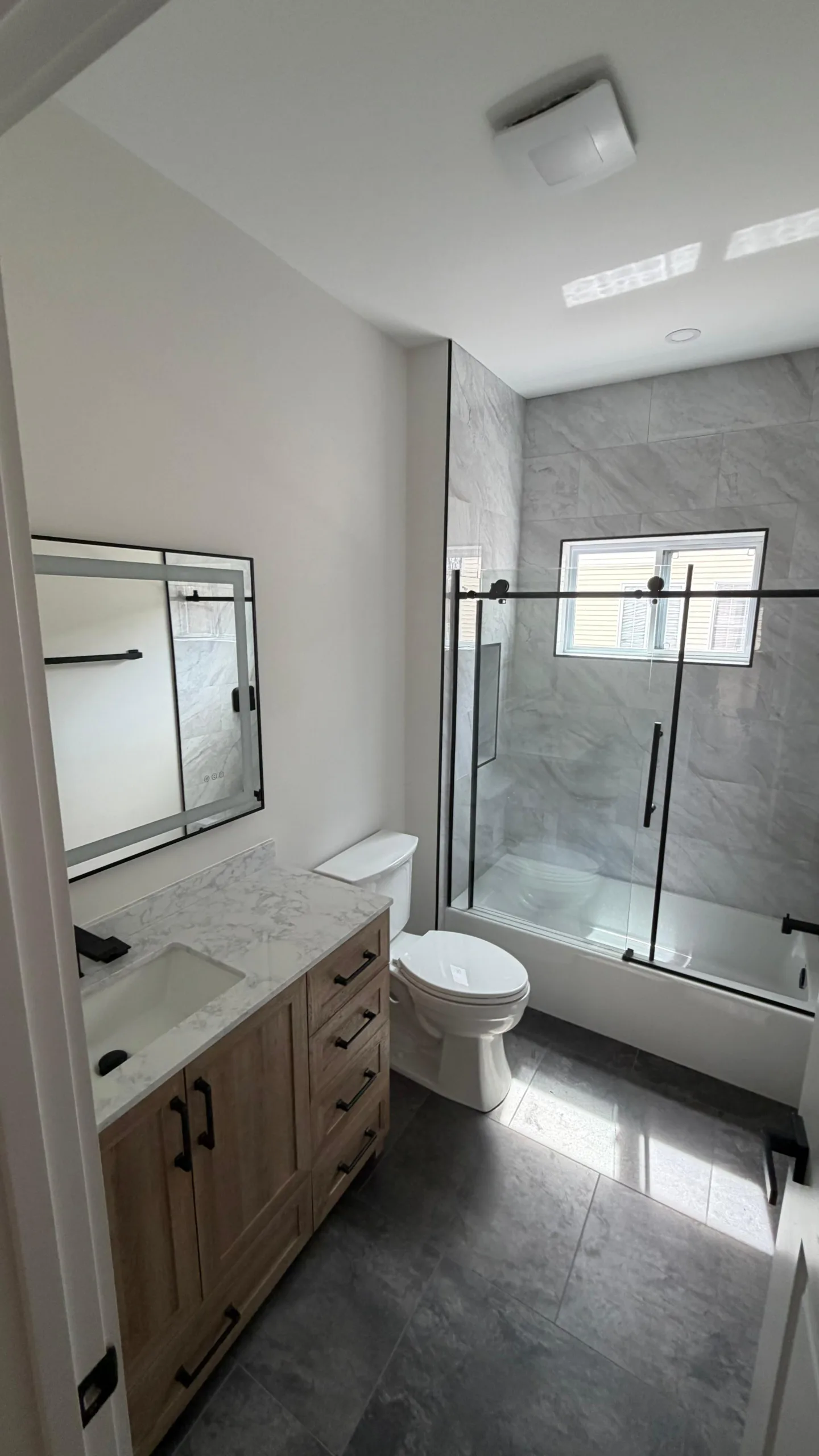 View from the doorway of the complete bathroom remodel, showing a new light wood vanity, a glass shower enclosure, and dark grey tile flooring.
